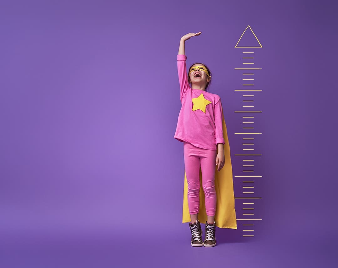 young girl in superhero costume stretching her arm above her head to measure her growth against a ruler on a bright purple wall